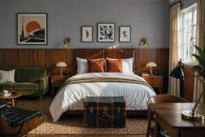 Hotel bedroom with dark wood panelled walls, leather headboard, brass wall lights, green velvet sofa, and vintage trunk at the foot of a white-dressed bed.