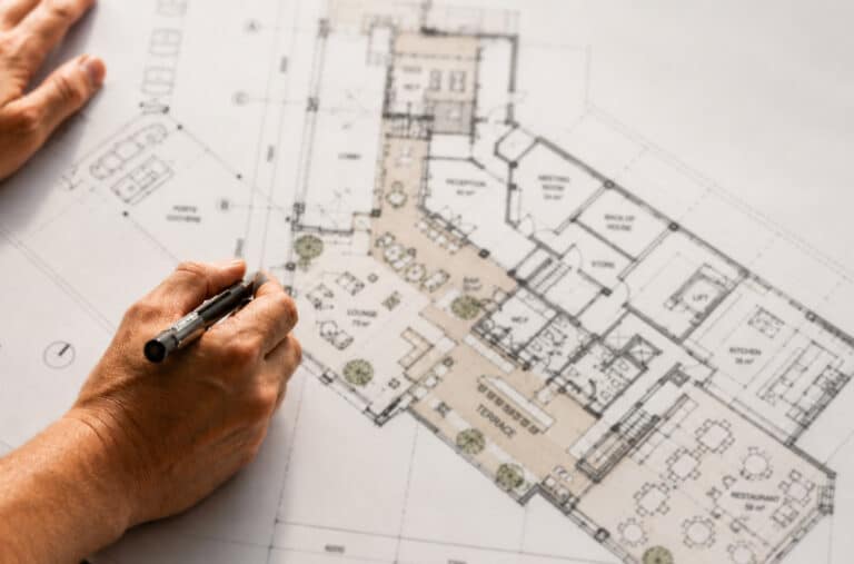 Close-up of a designer's hands holding a pen over a detailed architectural floor plan for a hotel, featuring labelled spaces including reception, lounge, bar, terrace, restaurant, kitchen, meeting room and back of house